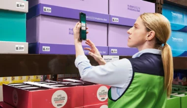 Une employée de magasin ou d'entrepôt, vêtue d'un polo bleu clair et d'une veste sans manches verte fluo, utilise un smartphone pour scanner un code-barres sur une boîte violette.