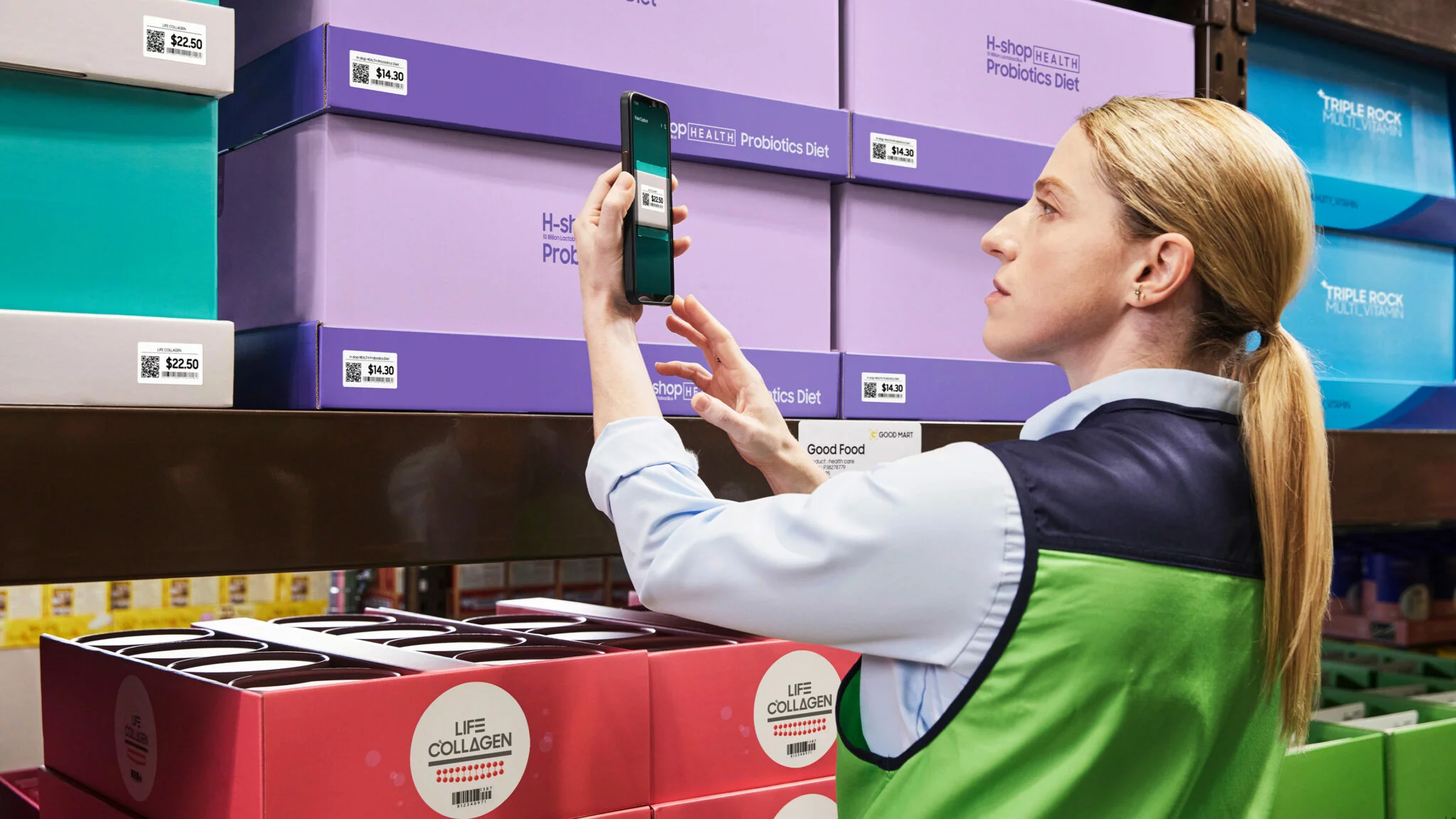 Une employée de magasin ou d'entrepôt, vêtue d'un polo bleu clair et d'une veste sans manches verte fluo, utilise un smartphone pour scanner un code-barres sur une boîte violette.