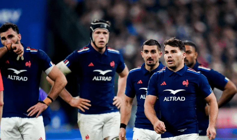 Plan moyen de plusieurs joueurs de l'équipe de France de rugby (XV de France) portant leurs maillots bleus officiels. Ils sont debout sur le terrain, le visage concentré et déterminé, en pleine action de jeu ou lors d'un arrêt de jeu.