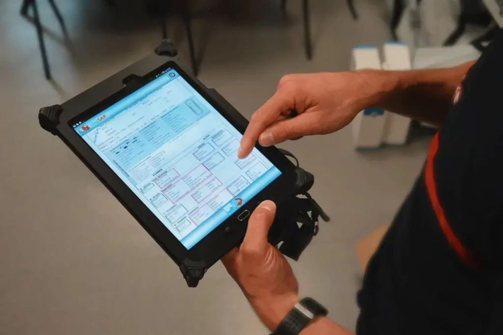 Gros plan sur les mains d'un secouriste ou d'un pompier, portant un uniforme bleu marine à rayures rouges, tenant une tablette numérique robuste.