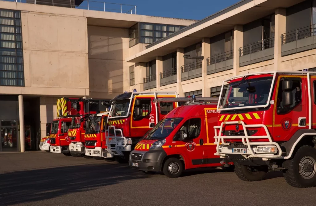 Une rangée de véhicules de sapeurs-pompiers rouges et blancs, dont des camions-citernes lourds et une fourgonnette plus petite avec l'inscription 'SAPEURS', est garée devant un bâtiment moderne à façade claire et des fenêtres sombres. Les véhicules sont vus de face, le soleil de l'après-midi éclairant la scène et projetant des ombres, mettant en valeur le caractère utilitaire et robuste de la flotte de secours.