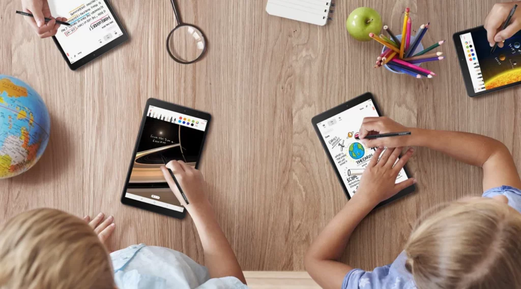 Vue en plongée sur un bureau en bois clair sur lequel sont disposés des fournitures scolaires et des tablettes. Deux enfants sont assis en bas, utilisant des tablettes.