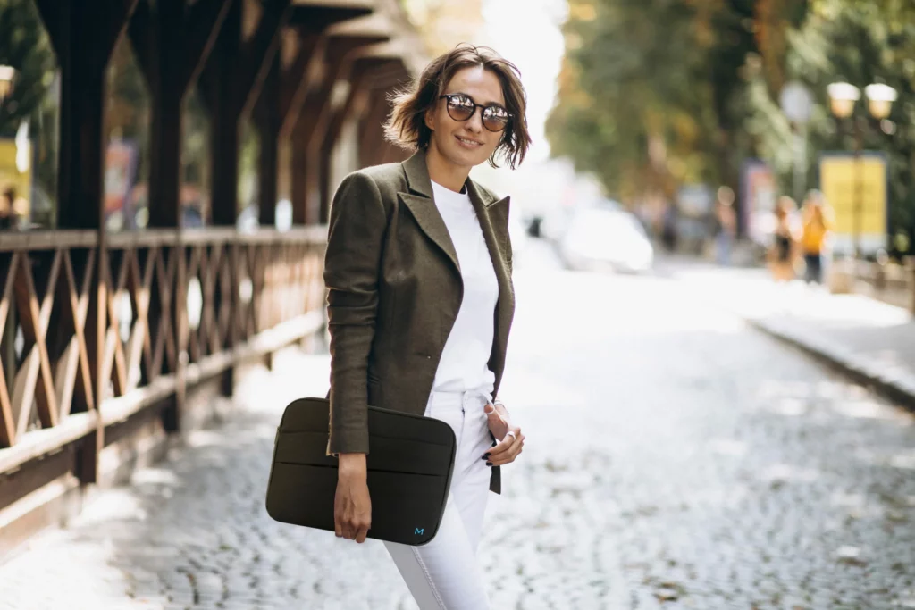 Plan moyen en extérieur d'une femme souriante, marchant dans une rue pavée bordée d'arbres, tout en tenant une housse d'ordinateur portable.