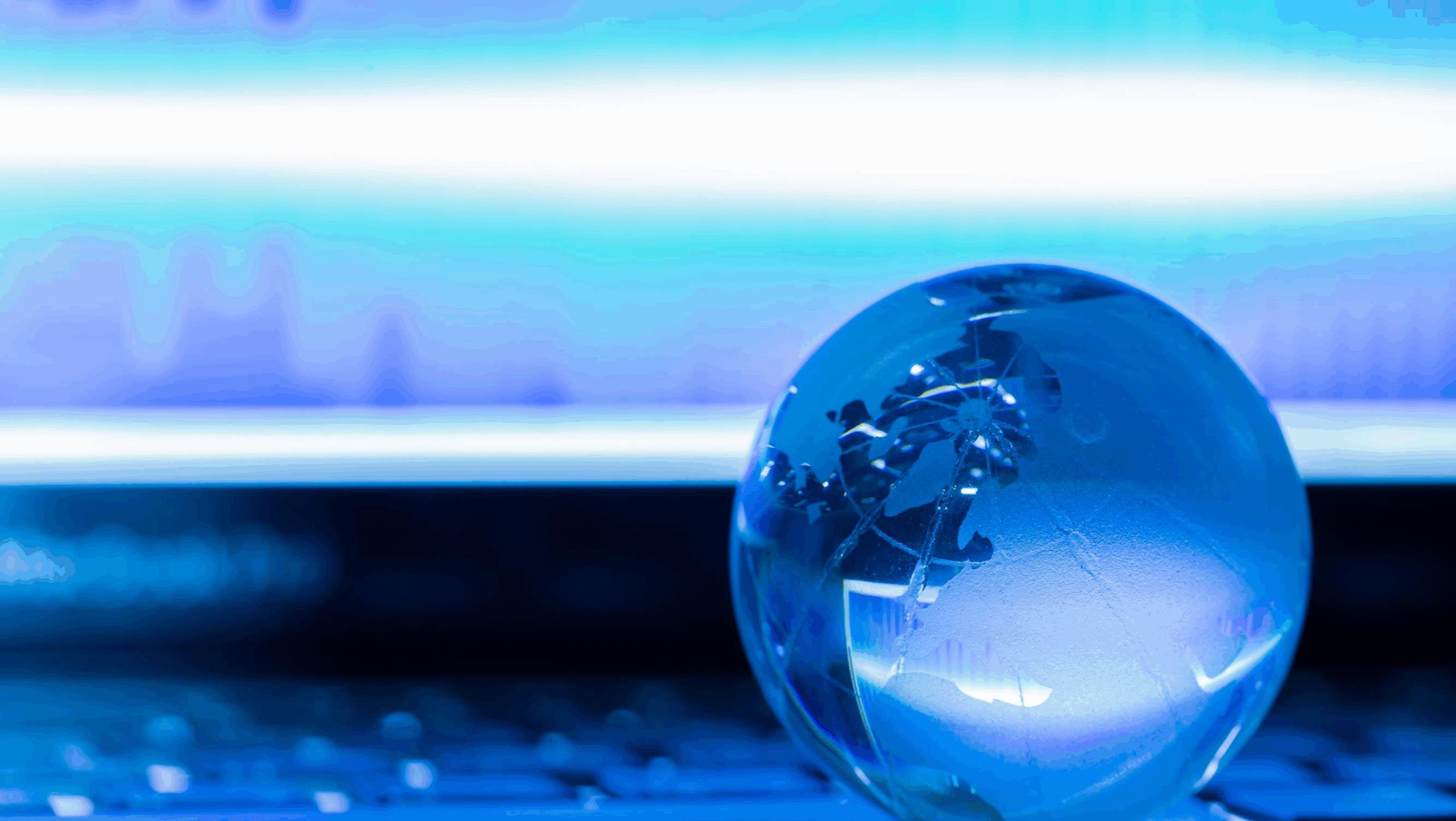 Un gros plan montre un globe terrestre miniature en verre bleu posé sur le clavier sombre d'un ordinateur portable. L'arrière-plan est dominé par une lumière bleue et blanche intense émanant de l'écran de l'ordinateur, créant une ambiance futuriste, technologique et globale. Le globe, qui représente des continents, est partiellement éclairé par la lumière vive.