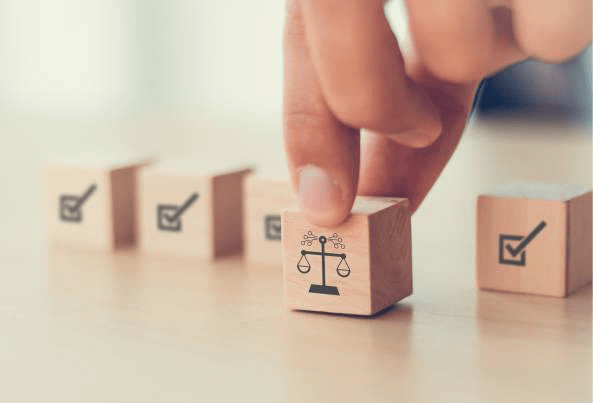 Image d'une main saisissant un petit cube en bois et le plaçant sur une table dans une rangée d'autres cubes. Le cube manipulé présente un symbole de balance de la justice (symbole du droit/légal) gravé sur une face. Les autres cubes visibles dans la rangée ont un symbole de coche ou de marque de validation noire gravé sur une face.