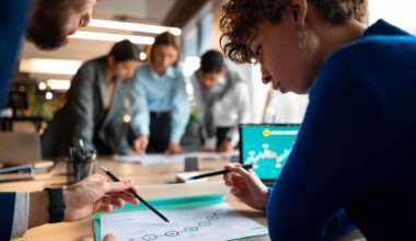 Une femme vêtue d'un pull bleu royal est penchée sur un graphique ou un document avec un homme dont la main est partiellement visible, pointant la feuille avec un stylo. Ils sont tous deux concentrés sur le document qui présente une série de cercles connectés sur une ligne. En arrière-plan, trois autres personnes sont penchées autour d'une autre table, et un ordinateur portable affiche un graphique sur son écran.