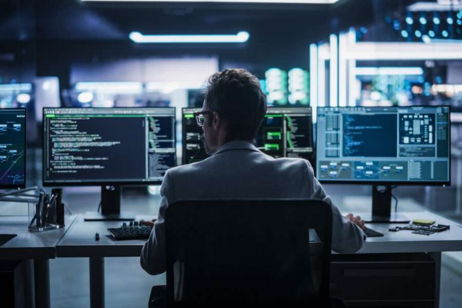 Vue de dos d'un jeune ingénieur travaillant sur un ordinateur dans un environnement de bureau technologique. Programmeur masculin écrivant du code logiciel pour un projet Blockchain, développant un système de backend (ou dorsal).