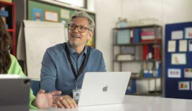 Une personne d'âge moyen avec des lunettes, portant une chemise bleue, est assis à une table dans une salle de classe, souriant et regardant vers le haut, peut-être en conversation. Un ordinateur portable Apple MacBook est ouvert devant lui, et une personne (partiellement visible sur la gauche) interagit avec lui, utilisant peut-être une tablette Apple iPad. La scène montre l'utilisation de la technologie Apple dans un contexte éducatif.
