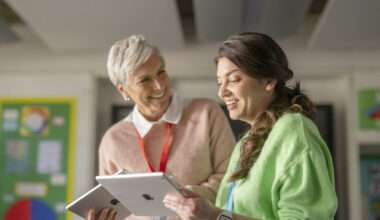 Deux enseignantes souriantes et complices dans une salle de classe moderne, utilisant des iPads pour des activités éducatives. La première femme, plus âgée, aux cheveux courts gris, porte un pull rose clair et sourit à la seconde femme, plus jeune, aux cheveux bruns, qui porte un pull vert citron et tient un iPad. L'image met en avant l'utilisation de la technologie (iPad) dans l'éducation.