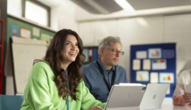 Femme souriante utilisant un iPad et un MacBook dans un environnement de bureau moderne ou une salle de classe. Un collègue plus âgé est assis à côté d'elle, travaillant également sur un ordinateur. Illustration d'une collaboration ou d'une formation professionnelle avec des produits Apple (iPad et MacBook).