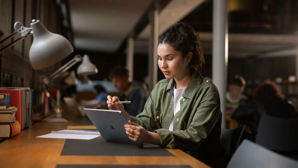 Étudiante écrivant sur un iPad avec un Apple Pencil la nuit dans une bibliothèque universitaire.