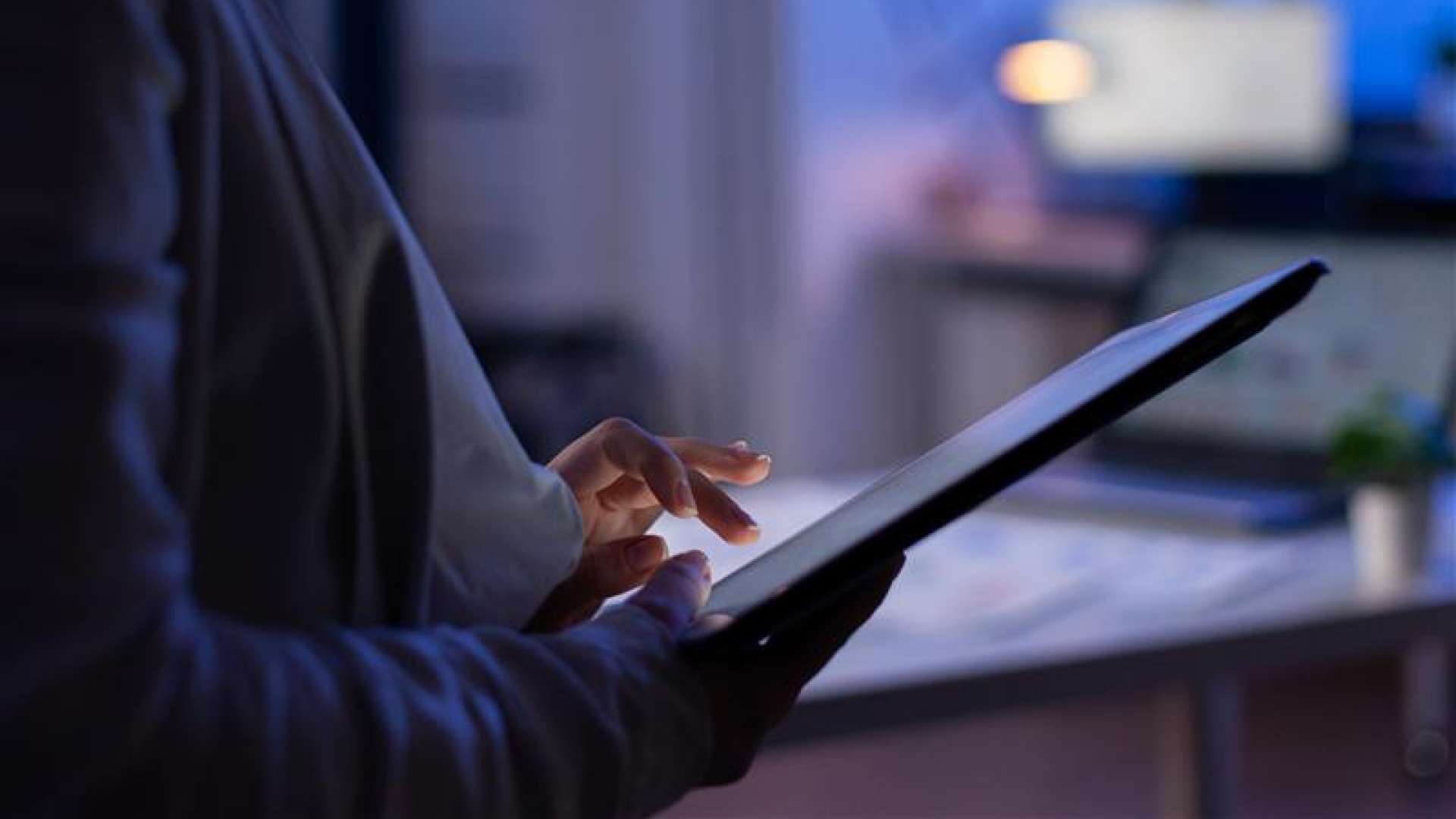 Gros plan sur les mains d'une personne utilisant une tablette tactile dans la pénombre, l'écran émettant une lumière bleue.