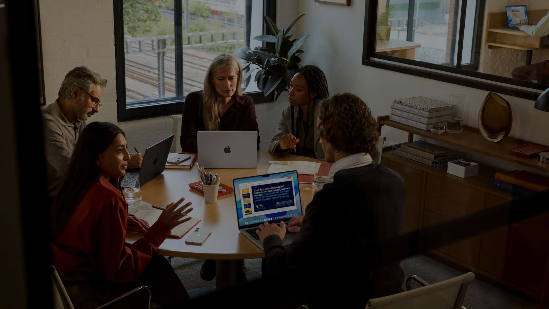 Un groupe de collègues de divers horizons est réuni autour d'une table ronde dans un bureau lumineux pour une session de travail. Ils utilisent plusieurs ordinateurs portables MacBook et prennent des notes.