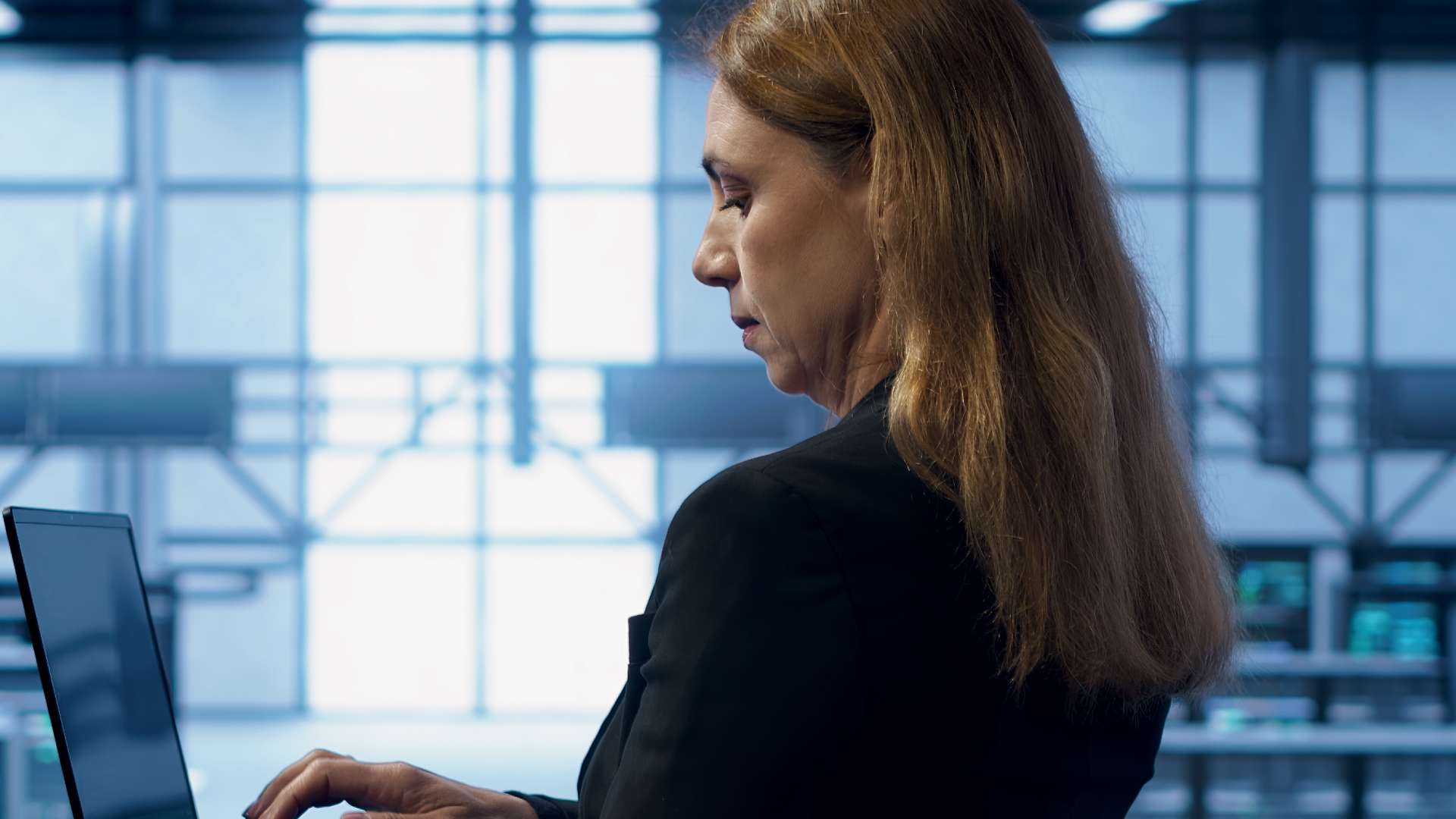 Une femme de profil, vêtue d'une veste noire, travaille avec concentration sur un ordinateur portable. Elle se trouve dans un vaste espace de travail moderne ou un centre de données aux tons bleutés, avec de grandes structures vitrées et des écrans de contrôle en arrière-plan.