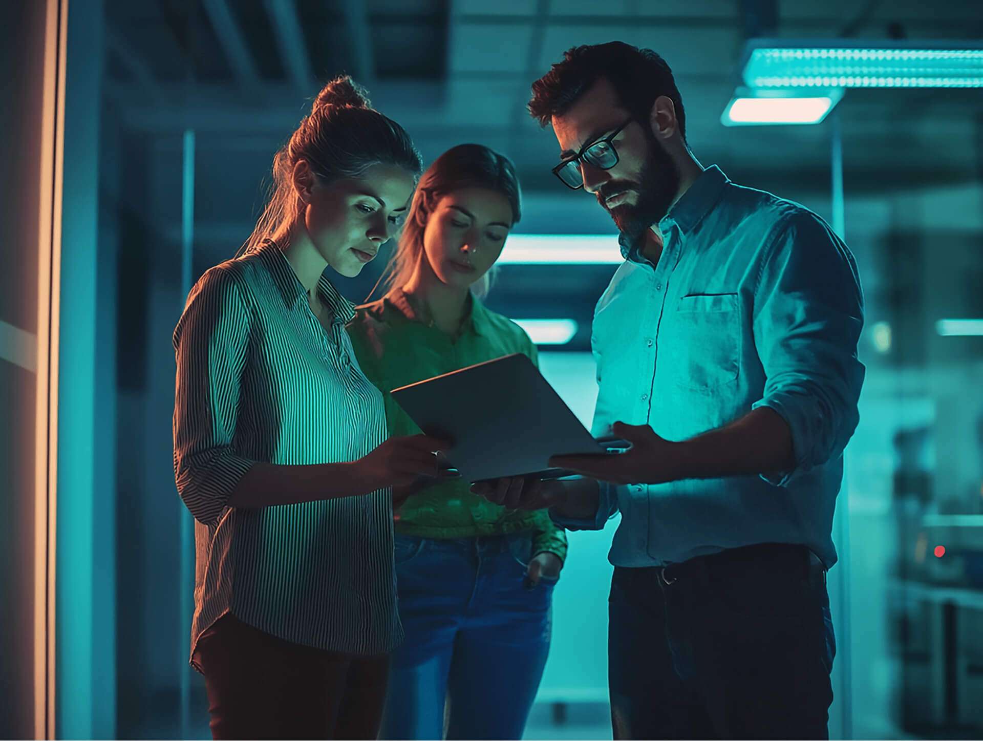 Trois collègues (deux femmes et un homme barbu portant des lunettes) sont regroupés dans un couloir de bureau sombre, éclairé par une lumière néon bleue. L'homme au centre tient une tablette ou un ordinateur portable, et les deux femmes regardent l'écran avec une expression concentrée. L'une des femmes est sur la gauche, vêtue d'une chemise rayée, et l'autre est à l'arrière-plan, vêtue d'une chemise verte.