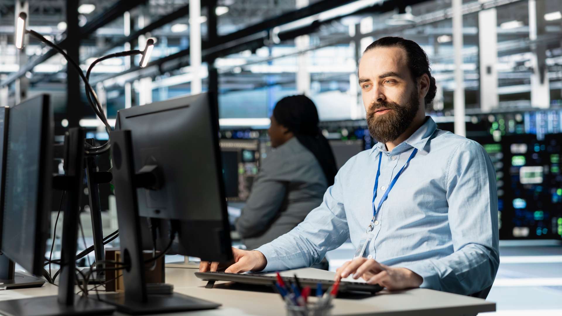 Un homme avec une barbe, portant une chemise bleue et un badge, travaille sur un ordinateur dans une salle informatique moderne et lumineuse. À l'arrière-plan, on aperçoit une autre personne travaillant devant des écrans de contrôle et des serveurs informatiques, illustrant un environnement de supervision technique et de sécurité.