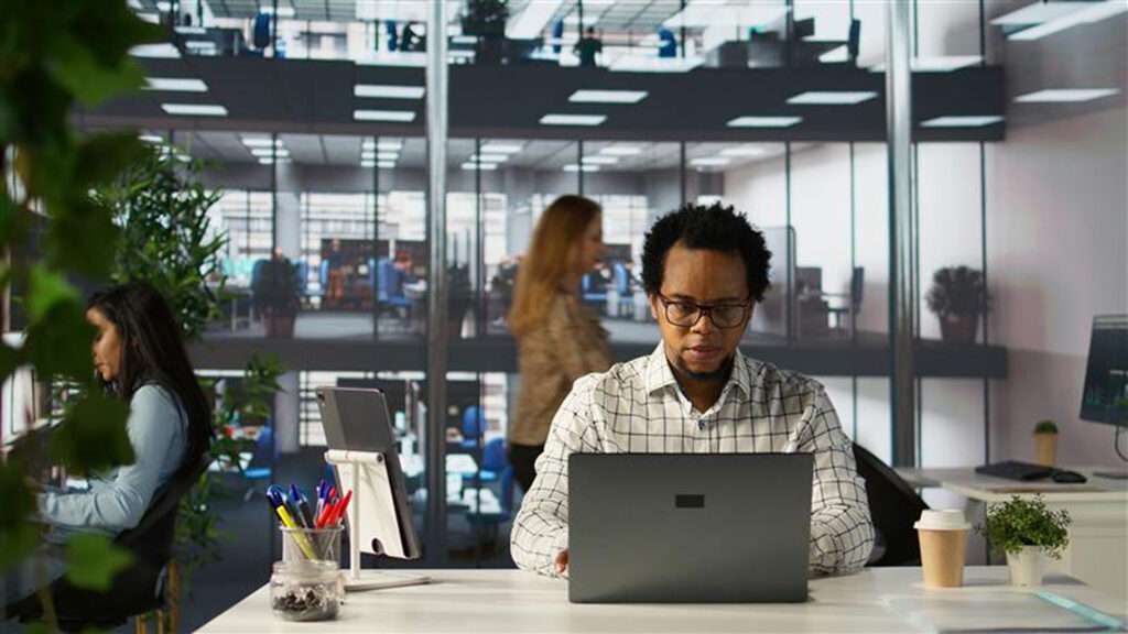 Vue de face d'un homme à lunettes travaillant sur son ordinateur portable dans un bureau moderne en open space. Il est installé à un bureau blanc bien organisé avec une tablette sur support, des fournitures et un café à emporter. À l'arrière-plan, d'autres collègues sont visibles dans un environnement de travail spacieux sur plusieurs niveaux, caractérisé par de grandes baies vitrées et des plantes vertes qui apportent une touche naturelle à l'espace professionnel.