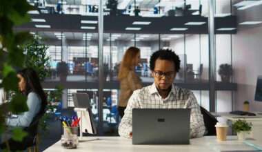 Vue de face d'un homme à lunettes travaillant sur son ordinateur portable dans un bureau moderne en open space. Il est installé à un bureau blanc bien organisé avec une tablette sur support, des fournitures et un café à emporter. À l'arrière-plan, d'autres collègues sont visibles dans un environnement de travail spacieux sur plusieurs niveaux, caractérisé par de grandes baies vitrées et des plantes vertes qui apportent une touche naturelle à l'espace professionnel.
