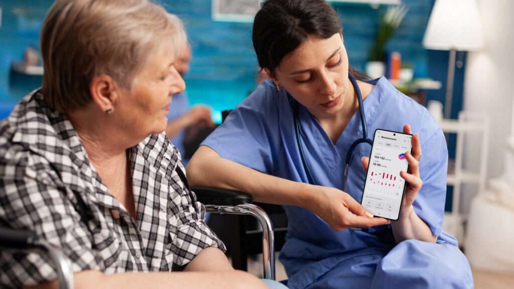 Une professionnelle de la santé en tenue bleue montre des données médicales, notamment un graphique de tension artérielle, sur l'écran d'un smartphone à une patiente âgée assise qui regarde attentivement l'écran.