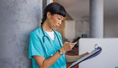 Une professionnelle de la santé en tenue médicale turquoise, portant un stéthoscope autour du cou, est appuyée contre un pilier gris. L'air concentré, elle consulte son smartphone d'une main tout en tenant un porte-bloc avec des documents de l'autre.