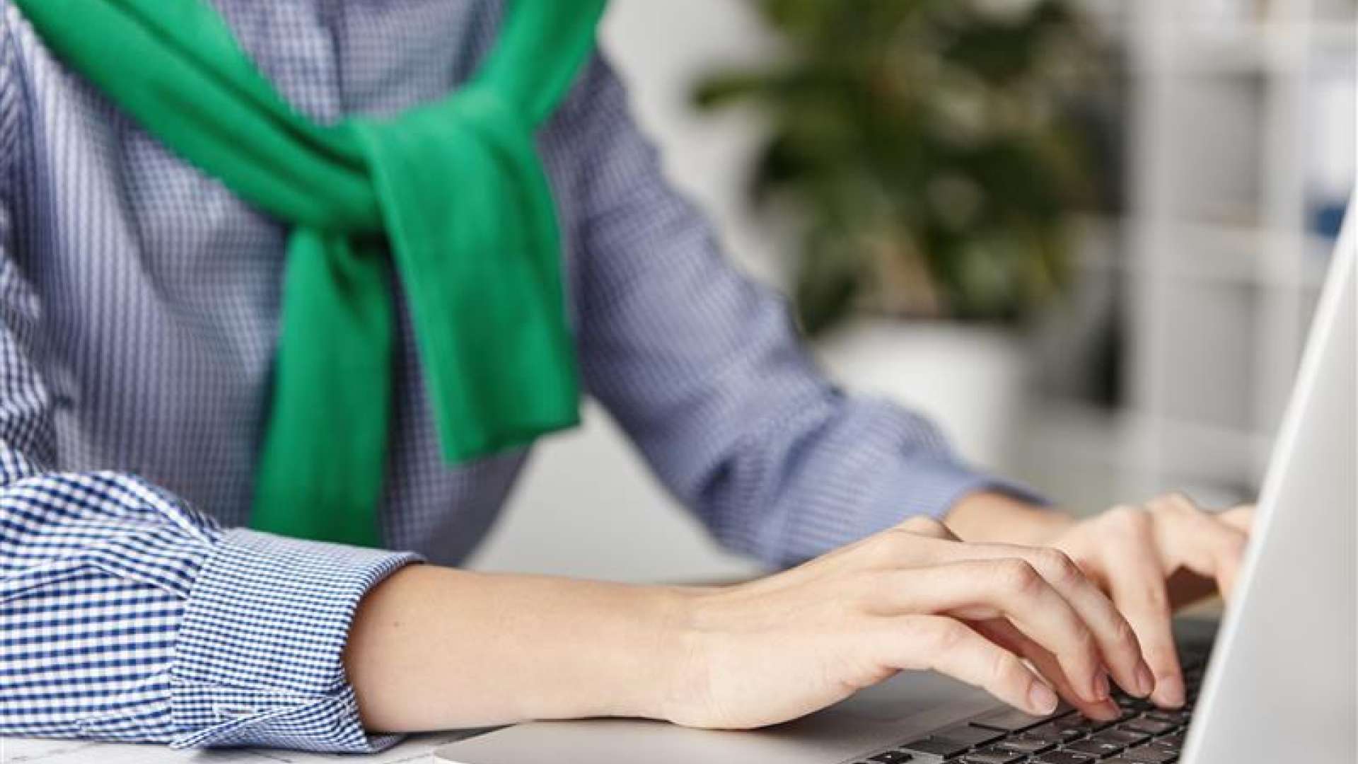 Plan rapproché sur les mains d'une personne en train de taper sur le clavier d'un ordinateur portable. La personne porte une chemise à petits carreaux bleus et blancs ainsi qu'un pull vert noué sur les épaules. L'arrière-plan est flou, laissant entrevoir un intérieur lumineux avec une plante verte.