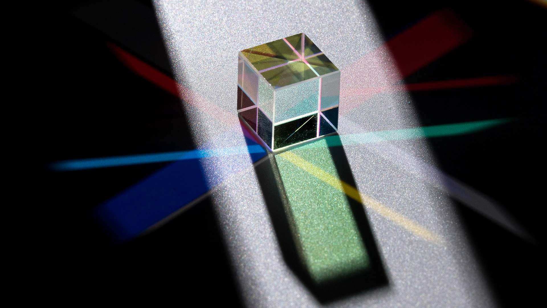 Un cube de verre dichroïque posé sur une surface métallique, décomposant la lumière en faisceaux colorés rouges, bleus et verts.