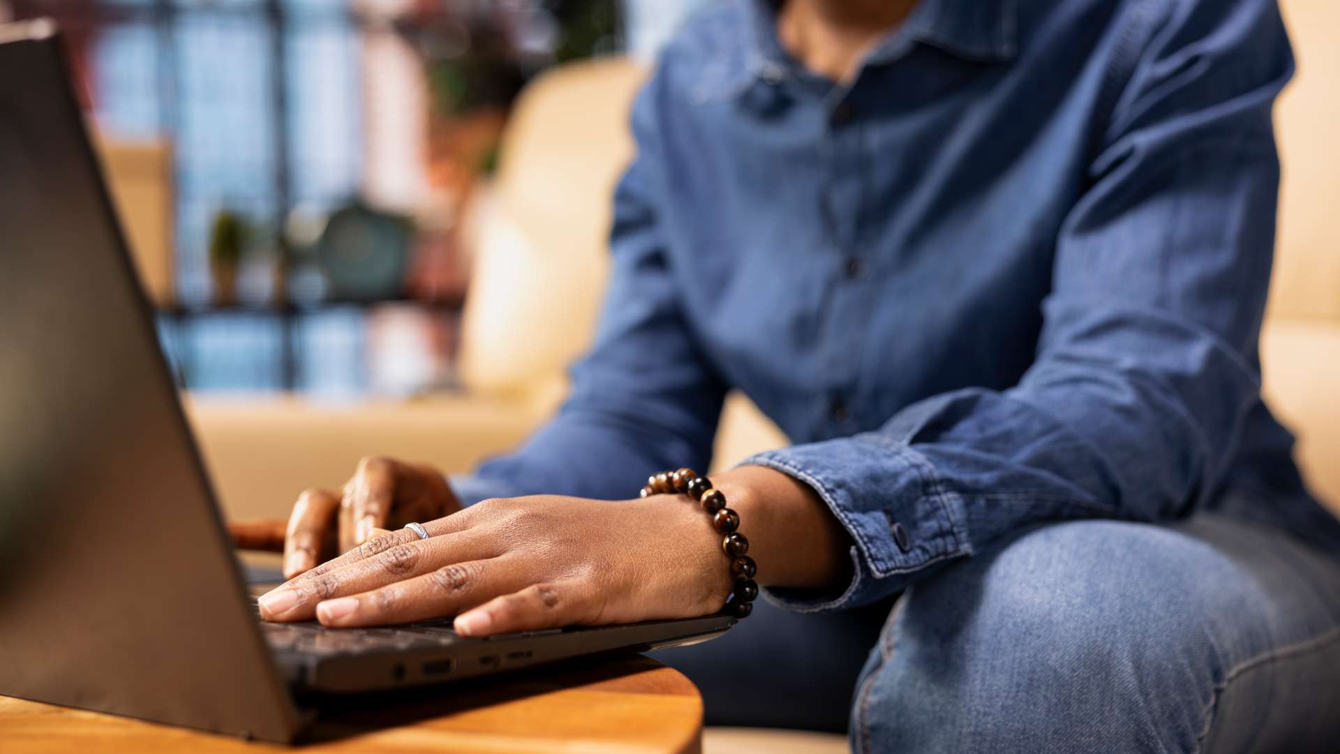 Gros plan sur les mains d'une personne en train d'utiliser un ordinateur portable posé sur une table basse en bois. La personne est vêtue d'une chemise en jean bleue et d'un pantalon en jean. Elle porte un bracelet en perles foncées au poignet droit et une bague à la main gauche. L'arrière-plan, flou, suggère un intérieur chaleureux avec ce qui semble être un canapé.