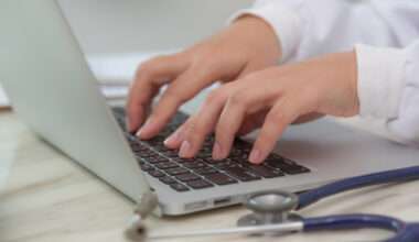 > Professionnel de santé utilisant un ordinateur portable pour saisir des données médicales avec stéthoscope posé sur le bureau