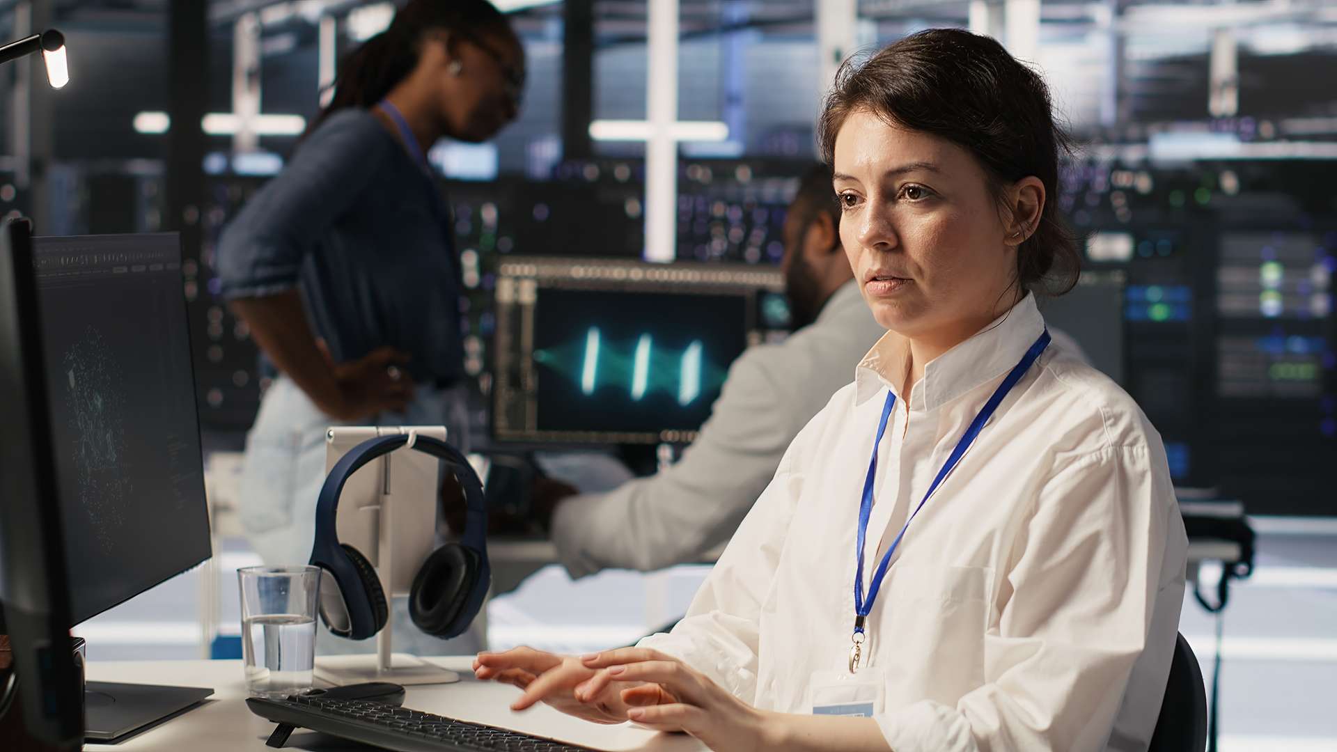 Technicien informatique travaillant sur ordinateur dans un data center avec écrans de monitoring et serveurs