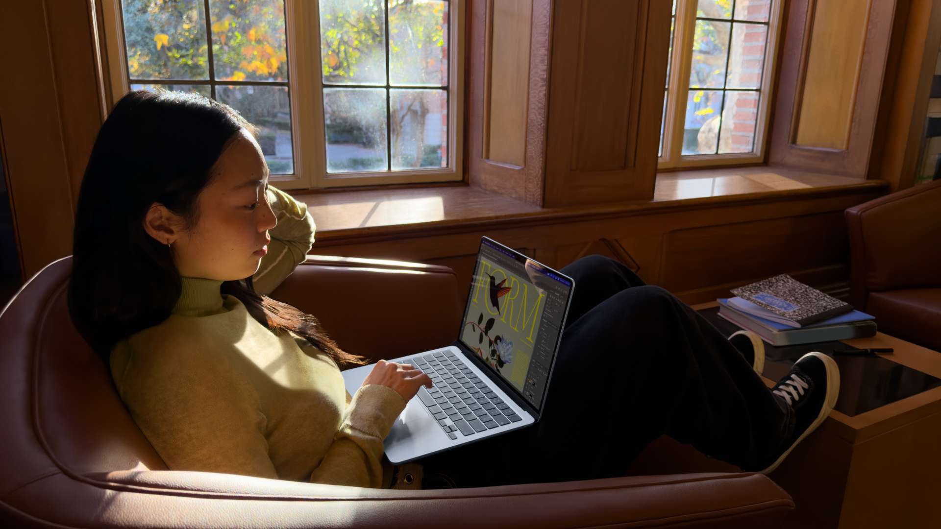 Personne assise dans un fauteuil près d’une fenêtre laissant entrer la lumière du jour, utilisant un MacBook Neo posé sur les genoux dans un intérieur chaleureux, avec des livres posés à proximité.