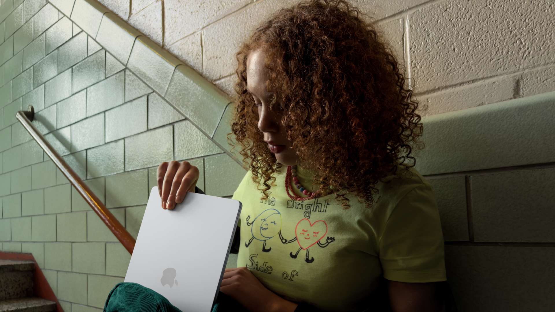 Personne assise dans un escalier intérieur avec des murs carrelés, tenant un MacBook Neo dans les mains et regardant l’écran, à proximité d’une rambarde et de marches, dans un environnement scolaire ou collectif.