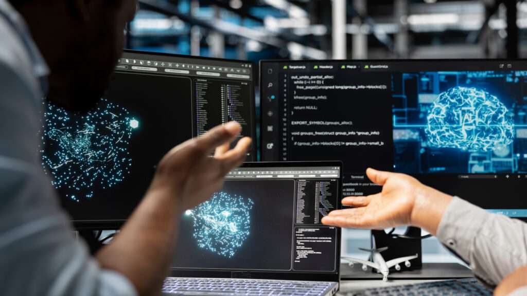 Poste de travail informatique avec plusieurs écrans affichant du code et des visualisations de données en forme de réseaux lumineux, tandis que deux personnes échangent en pointant les écrans dans un environnement technologique.
