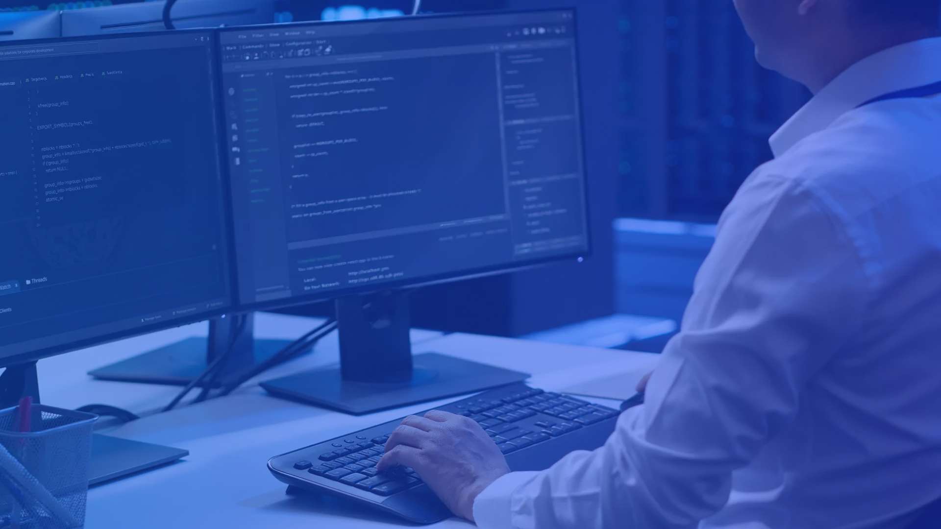 Personne travaillant sur un ordinateur avec deux écrans affichant du code informatique dans un environnement de bureau, avec un filtre bleu dominant et les mains posées sur un clavier.