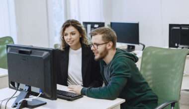 Deux personnes assises côte à côte dans un bureau, travaillant sur un ordinateur de bureau, en train de consulter le même écran avec clavier et souris, dans un environnement de travail moderne avec d’autres postes informatiques en arrière-plan.