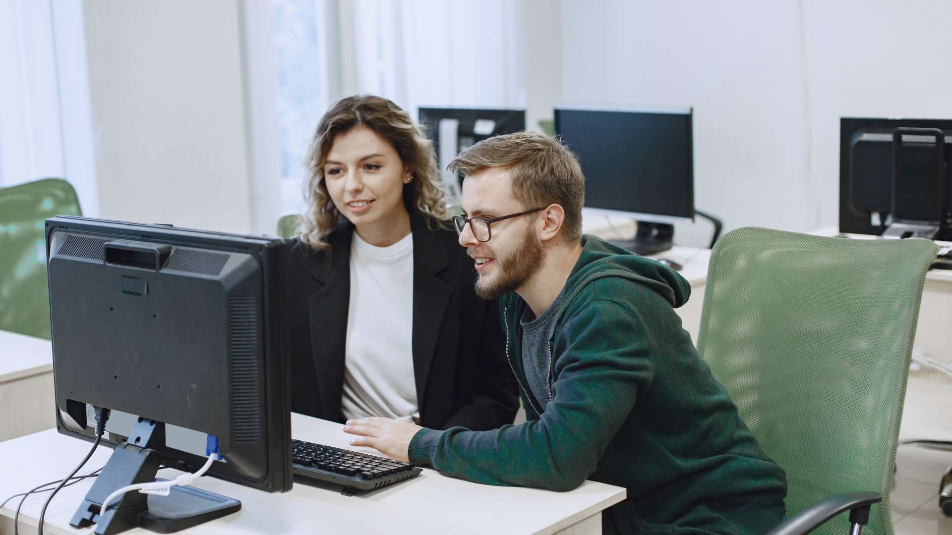 Deux personnes assises côte à côte dans un bureau, travaillant sur un ordinateur de bureau, en train de consulter le même écran avec clavier et souris, dans un environnement de travail moderne avec d’autres postes informatiques en arrière-plan.