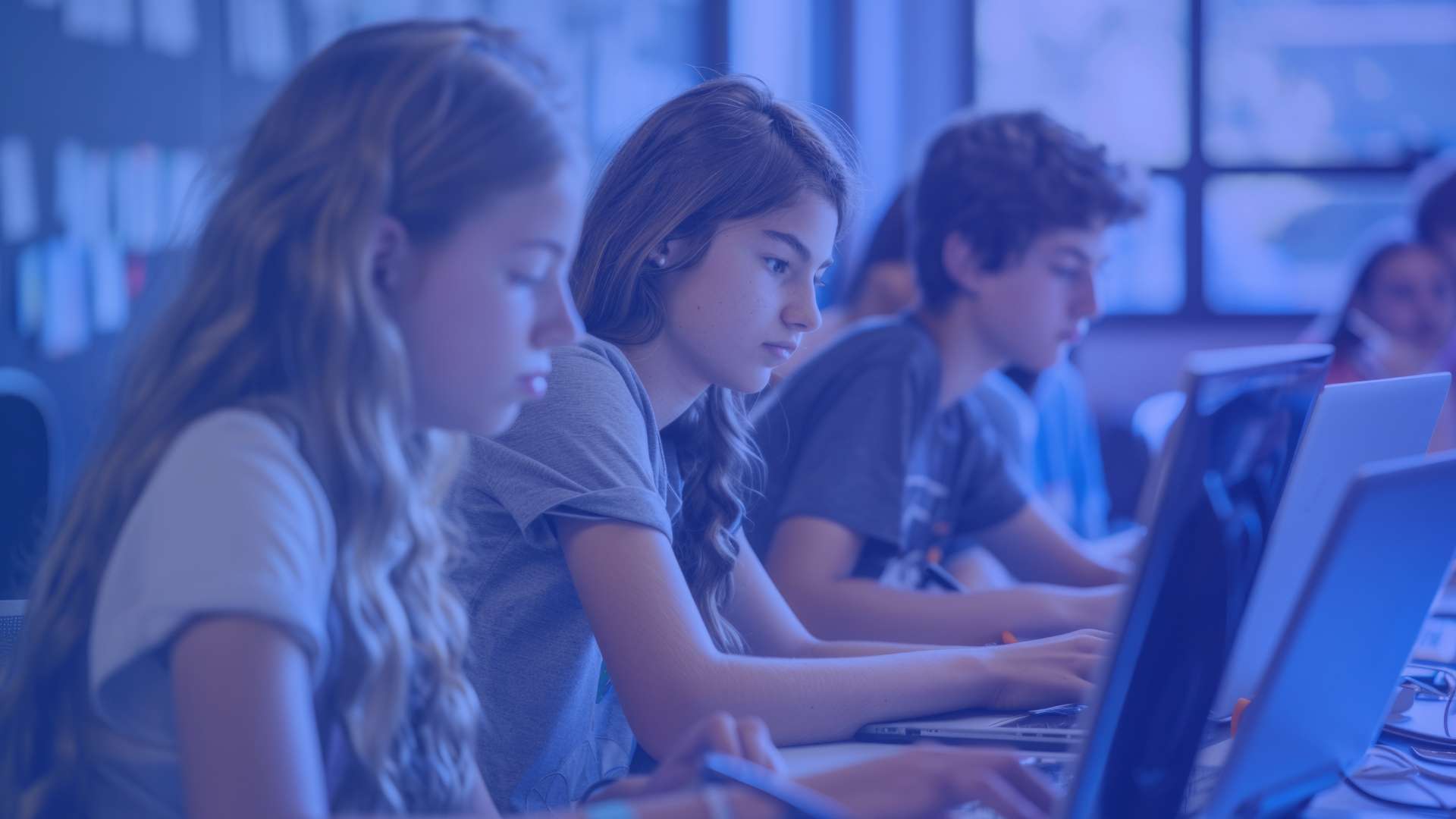 Salle de classe où plusieurs élèves sont assis en rangée et travaillent chacun sur un ordinateur portable, concentrés sur leurs écrans, avec une teinte bleutée appliquée à l’image et des bureaux alignés.