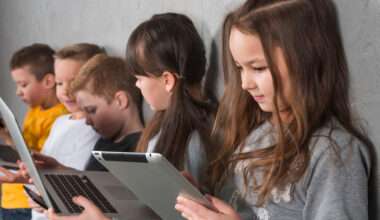 Groupe d’enfants assis côte à côte contre un mur, chacun utilisant un appareil numérique (tablette ou ordinateur portable), concentrés sur leurs écrans dans un contexte d’apprentissage ou de loisir.