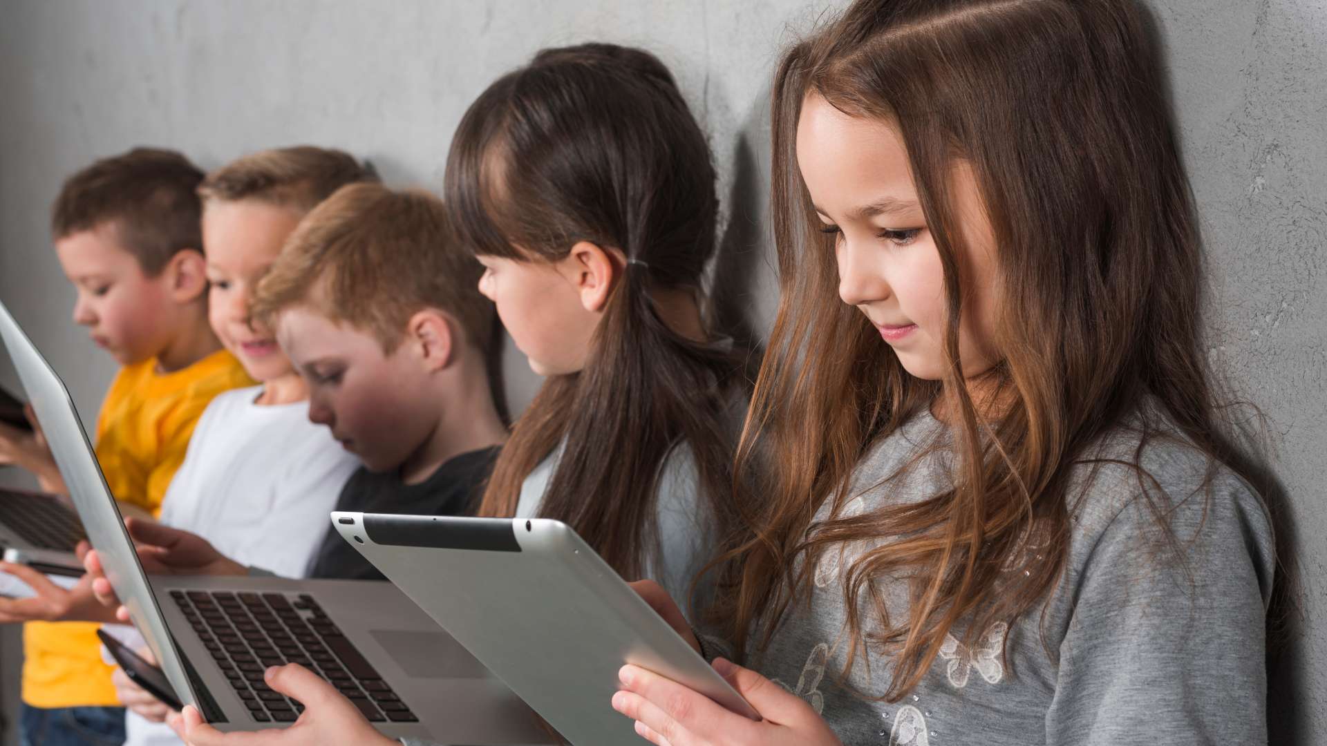Groupe d’enfants assis côte à côte contre un mur, chacun utilisant un appareil numérique (tablette ou ordinateur portable), concentrés sur leurs écrans dans un contexte d’apprentissage ou de loisir.