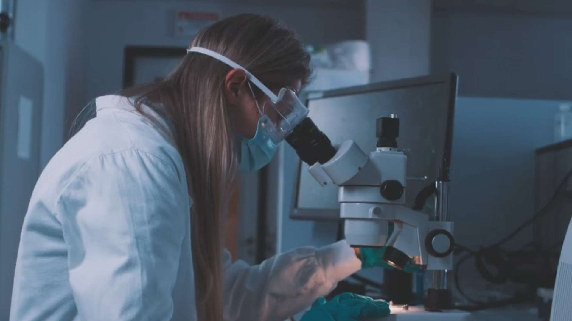 Personne en blouse de laboratoire portant des gants et des lunettes de protection, observant un échantillon sous un microscope dans un environnement scientifique.