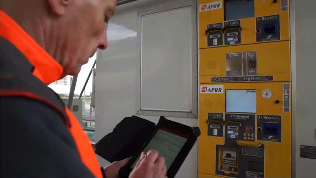 Personne portant un gilet haute visibilité utilisant une tablette tactile devant une borne de péage automatique APRR. La borne jaune comporte un écran, un lecteur de carte bancaire, un distributeur de tickets et un emplacement pour le reçu. La scène se déroule dans un environnement technique avec des équipements de péage visibles à l’arrière-plan.