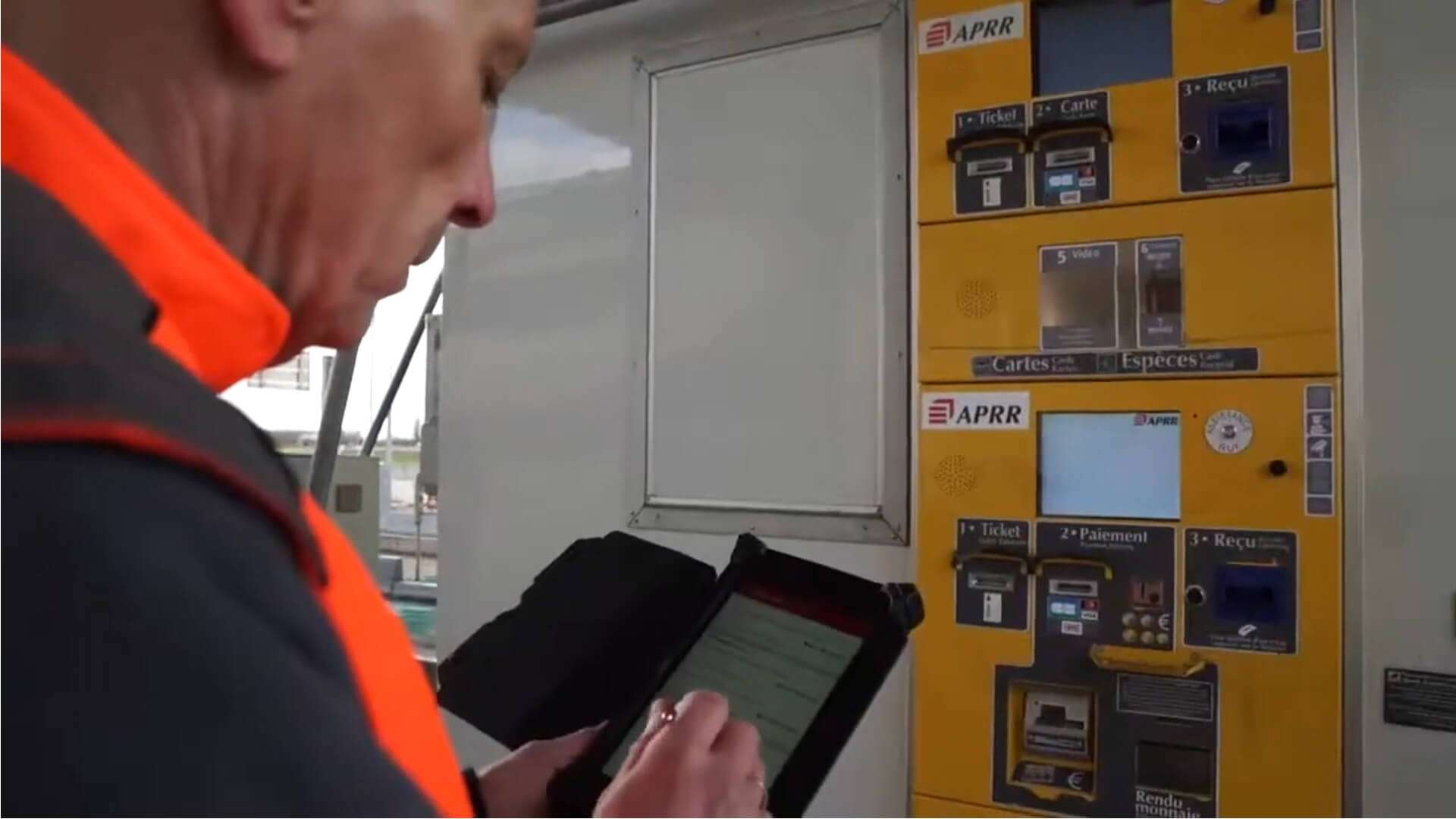 Personne portant un gilet haute visibilité utilisant une tablette tactile devant une borne de péage automatique APRR. La borne jaune comporte un écran, un lecteur de carte bancaire, un distributeur de tickets et un emplacement pour le reçu. La scène se déroule dans un environnement technique avec des équipements de péage visibles à l’arrière-plan.