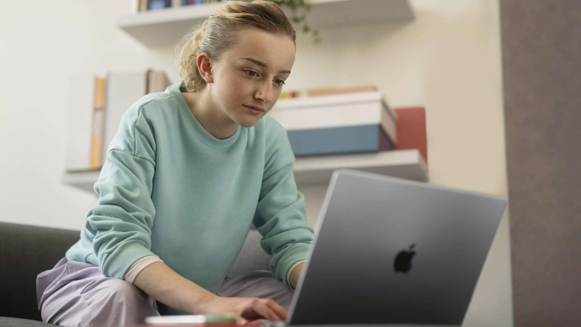 Une personne est assise sur un canapé et utilise un ordinateur portable dans un espace intérieur. Des étagères avec des livres et des objets de rangement sont visibles à l’arrière-plan. La scène est éclairée par une lumière douce et met en avant l’utilisation d’un appareil numérique dans un environnement calme et domestique.