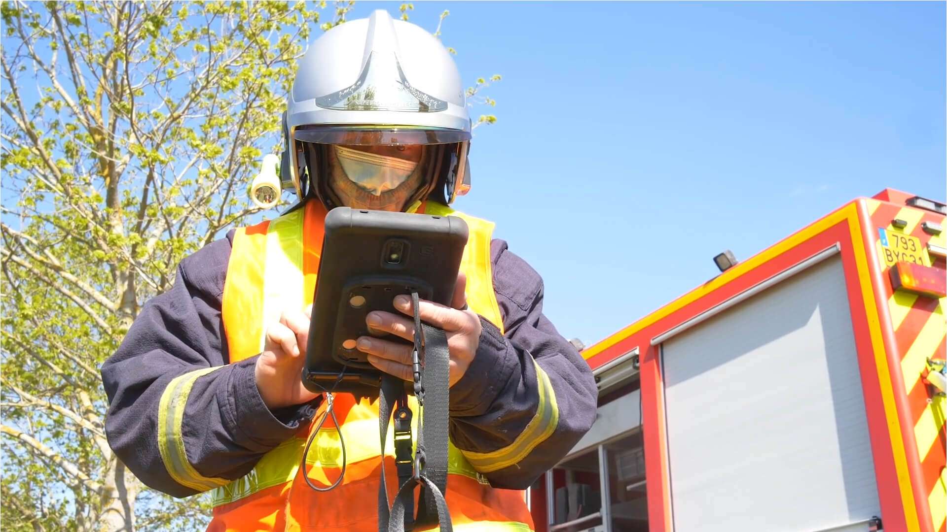 Personne équipée d’un casque, d’un masque de protection et d’un gilet haute visibilité utilisant une tablette numérique à l’extérieur. Un véhicule d’intervention rouge et jaune est stationné à proximité. La scène se déroule en plein jour, sous un ciel dégagé.