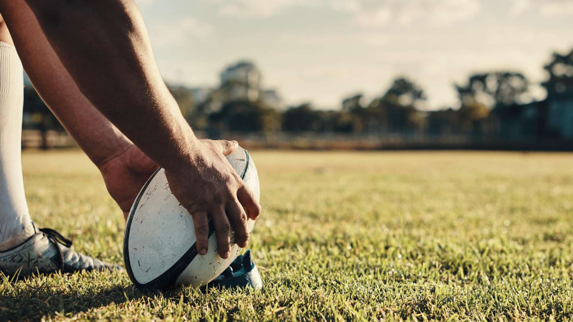 Gros plan sur une personne plaçant un ballon de rugby sur un tee au milieu d’un terrain en herbe. Seules les jambes, les chaussures et les mains sont visibles. L’arrière-plan est flou, avec des arbres et un ciel lumineux en lumière naturelle.